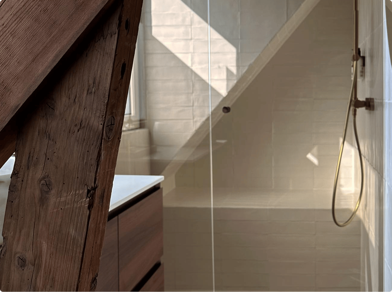 Charaktervolles x²o Bad mit schräger Decke und sichtbarer Holzbalkenstruktur. Rechts eine Dusche mit weißen Fliesen und goldfarbiger Regendusche. Links ist ein Holzbadmöbel unter einem Fenster mit einfallendem Sonnenlicht zu sehen.